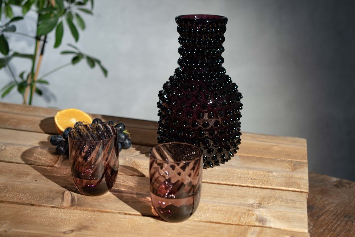 Violet Hobnail Carafe on a wooden pallet surrounded by a cut orange and glasses, with a green plant and a wall in the background 